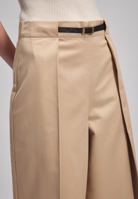 Pantalon beige taille haute avec une coupe ajustée, comportant un pli latéral, une ceinture noire et des détails en doré. Tissu à texture lisse.