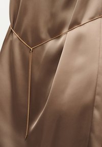 Λεπτή ζώνη με χρυσή αλυσίδα και μικρό κυκλικό κούμπωμα, τοποθετημένη πάνω σε λεία, καφέ σατέν ύφασμα.