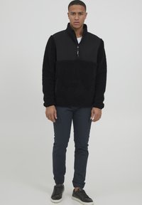Schwarzer Fleece-Pullover mit einem halben Reißverschluss-Kragen, Fronttasche und einem glatten oberen Bereich. Kombiniert mit dunklen Hosen und schwarzen Turnschuhen.