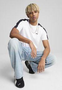 Camiseta blanca con detalles negros, vaqueros anchos azules y zapatillas negras. Cadenas en el cuello, con un peinado texturizado y varios anillos.