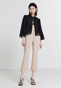 Donna che indossa una maglietta nera con bottoni e rifiniture in pizzo sopra pantaloni beige a vita alta e tacchi neri con cinturini, in piedi su uno sfondo bianco semplice.