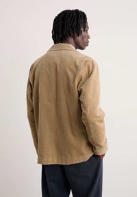 Gaiši brūns korduroja apakšžakets ar klasisku apkakli un pogu priekšpusē, ar brīvu piegriezumu un redzamu ribotu tekstūru.