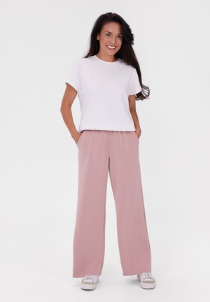 Frau steht mit Händen in den Taschen, trägt weißes T-Shirt, pinke, weit geschnittene Hose und weiße Sneaker vor einfarbigem Hintergrund.