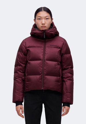 Personne portant une doudoune rouge foncé avec un col haut et une capuche, debout devant un fond gris clair uni.