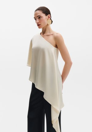 Donna che indossa un top asimmetrico crema monospalla con pantaloni neri e grandi orecchini rotondi dorati, in piedi su sfondo bianco.