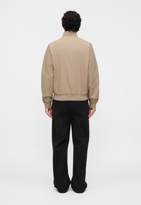 Άνδρας με σγουρά σκούρα μαλλιά, φορώντας μπεζ σακάκι και μαύρο παντελόνι, στέκεται με την πλάτη προς τα εμπρός σε ένα απλό λευκό φόντο.