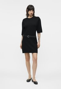 Femme debout vêtue d'une robe noire à manches courtes avec une ceinture, des ballerines noires, et des tatouages visibles sur les bras, sur un fond blanc uni.