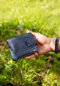 Gray fabric wallet with a textured surface, featuring a smooth black leather bottom and a snap closure. Metal brand logo is visible.