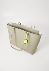 Beige-kangaskassi, jossa on kaksi kahvaa, teksturoitu materiaali ja hopeinen sisustus, jossa on hillitty logopainatus; mukana on keltainen yksityiskohtatag.