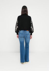 Suéter corto negro con bordado floral blanco en las mangas, combinado con jeans acampanados azules. Los jeans tienen bolsillos traseros y un lavado claro.