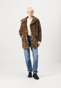 Leopardprint nepbontjas met een brede kraag, knoopsluiting en twee voorzakken. Gecombineerd met lichtblauwe jeans en zwarte schoenen.