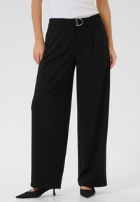 Pantalons larges noirs avec un devant plié, une boucle de ceinture argentée en forme de D et une texture de tissu lisse. Portés avec des talons aiguilles noirs.