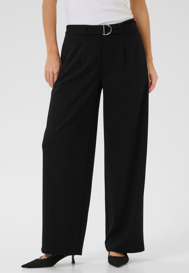 Pantalons larges noirs avec un devant plié, une boucle de ceinture argentée en forme de D et une texture de tissu lisse. Portés avec des talons aiguilles noirs.