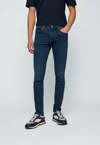 Jean slim-fit bleu marine avec une texture en denim lisse, design à cinq poches et détails en matériel argenté, associé à des baskets multicolores.