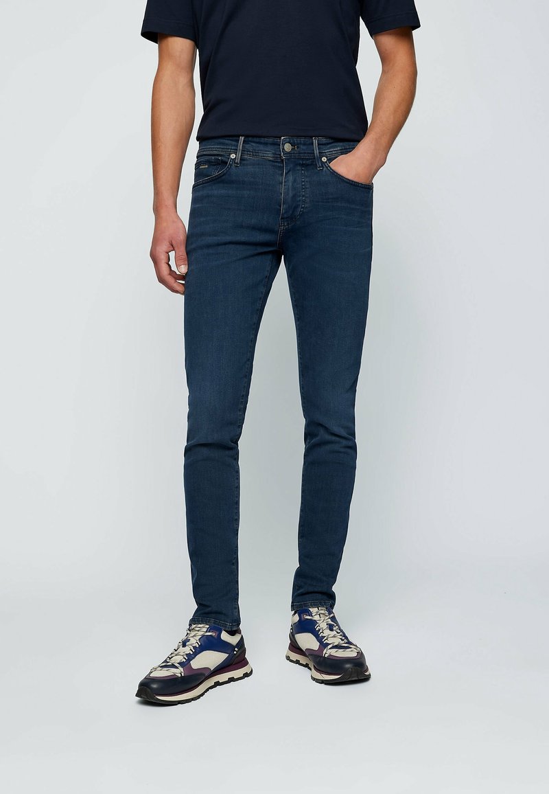 Jean slim-fit bleu marine avec une texture en denim lisse, design à cinq poches et détails en matériel argenté, associé à des baskets multicolores.