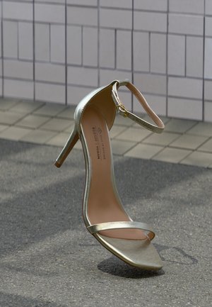 Goldene Sandalette mit hohem Absatz, Knöchelriemen, quadratischer Spitze und dünnem Absatz auf grauem Pflaster vor einer gefliesten Wand.