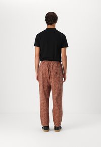 Brown patterned trousers with a relaxed fit, elastic waistband, and a small branded patch on the back pocket, paired with a black tee.