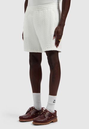 Donkerharig persoon die witte textuurige shorts draagt, witte sportsokken en bruine leren bootschoenen met dikke rubberen zolen.