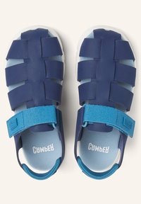 Siniläbised kummist sandaalid, millel on rihmadega disain. Valge tald, reguleeritav sinine rihm ja pehme sisetald on nähtavad. Logo on trükitud tallale.