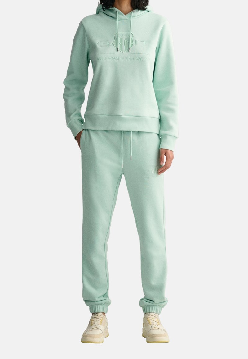 Persoon in een lichtgroene hoodie en bijpassende sweatpants met een geperst logo, gecombineerd met crèmekleurige sneakers.