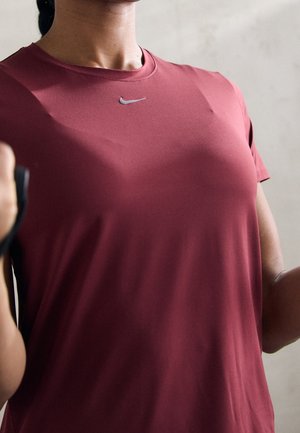 Gros plan d’une personne portant un t-shirt bordeaux Nike, tenant des bandes de résistance noires, en train de faire de l’exercice sur un fond uni.