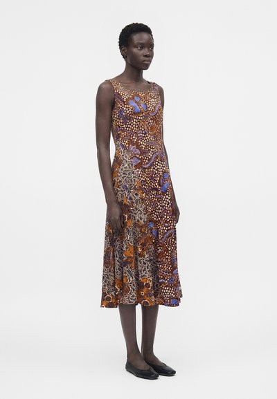 Γυναίκα που φοράει αμάνικο φόρεμα μήκους μέχρι τα μοσχάρια με καφέ, μπλε και πορτοκαλί αφηρημένα σχέδια, στέκεται μπροστά σε μια μονόχρωμη γκρι φόντο.