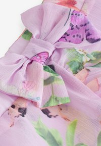 Lavendelblumenstoff mit einer Schleifendetail, der verschiedene bunte Blumen- und Schmetterlingsdrucke aufweist. Die Textur wirkt weich und leicht.