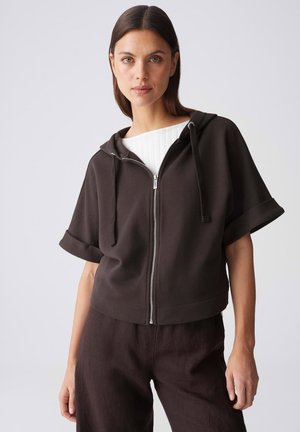 Donna con capelli castani lisci indossa una felpa con cappuccio a maniche corte marrone scuro con zip sopra una canotta bianca e pantaloni marroni scuri, in piedi davanti a uno sfondo semplice.