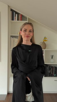 Zwarte lange mouwen jurk met een gewikkeld ontwerp en brede manchetten; heeft een glanzende textuur en subtiele plooien. Subtiele sieraden accentueren de look.