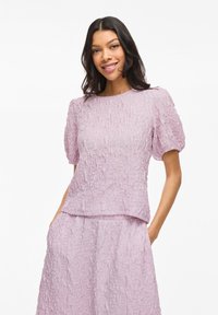 Top textured viola chiaro con maniche a sbuffo corte, caratterizzato da un collo rotondo e una vestibilità rilassata. Il tessuto ha un aspetto increspato e stratificato.