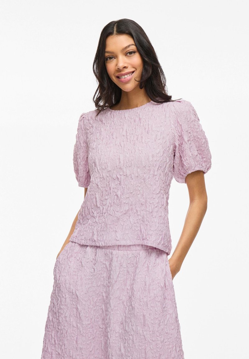 Top textured viola chiaro con maniche a sbuffo corte, caratterizzato da un collo rotondo e una vestibilità rilassata. Il tessuto ha un aspetto increspato e stratificato.