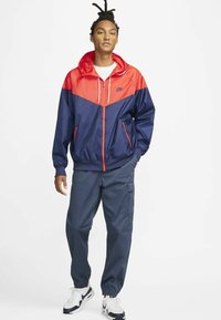 Nike vėjo apsiaustas, mėlynas ir raudonas, su užtrauktuku priekyje, reguliuojama kapuka ir priekinėmis kišenėmis. Suderintas su tamsiomis cargo kelnėmis ir sportbačiais.