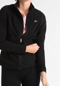 Persoon in een zwarte ritsjack over een roze shirt, die hun linkeronderarm vasthoudt met hun rechterhand.