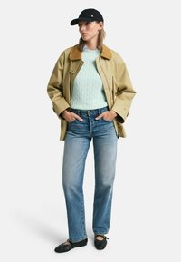 Giacca leggera beige con colletto marrone, maglione a trecce verde menta, jeans blu e ballerine nere intrecciate, completato da un cappellino nero.