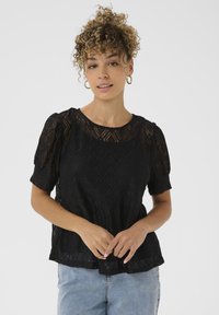 Top in pizzo nero con maniche corte a sbuffo, caratterizzato da un motivo a diamante e un collo rotondo. Indossato sopra jeans in denim azzurro chiaro.