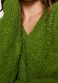 Pull à col en V vert vif, fabriqué en tissu doux et texturé avec une coupe ample, présentant un aspect tissé douillet.