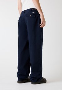 Marineblaue Hosen mit weitem Bein aus weichem Stoff, mit zwei aufgesetzten Gesäßtaschenklappen mit Knopfverschluss und einem Logodetail in der Nähe des Bunds.