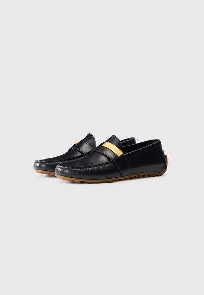 Svarta läderloafers med ett gyllene accentband, sömnadsdetaljer och en kontrasterande brun gummisula. Slät yta och rundad tådesign.
