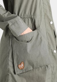 Ręka w kieszeni oliwkowej kurtki zapinanej na zatrzask, z małą haftowaną naszywką lamy na klapie kieszeni.