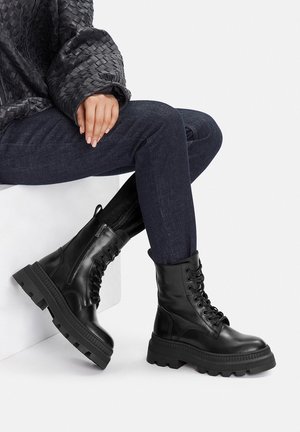 Schwarze Leder-Kampfstiefel mit Schnürdesign, klobiger Gummisohle und strukturierter Oberfläche, getragen zu dunklen Jeans und einem gemusterten Oberteil.