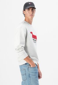 Light gray sweatshirt with red embroidered text, round neckline, and ribbed cuffs, paired with blue denim jeans and a dark cap.
