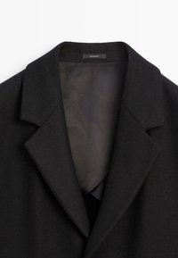 Cappotto di lana nera con colletto a punta, dotato di una fodera interna morbida e scura e di una superficie esterna con texture. Design semplice ed elegante.