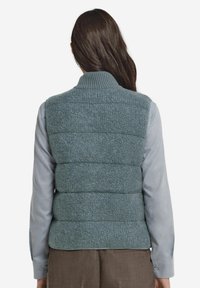 Texturerad blågrön puffväst med ribbad krage, vertikala sömmar och lätt isolering, lager-på-lager över en ljusgrå skjorta.