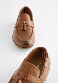 Mocassins en cuir marron avec un dessus texturé, des pompons décoratifs, des accents cousus et une doublure intérieure à motifs avec des rayures.