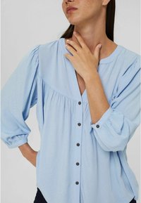 Blouse bleu clair en tissu texturé, avec un col en V, un design plissé, de longues manches bouffantes et de petites fermetures à boutons marron.