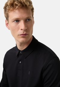 Schwarzes Poloshirt aus strukturiertem Stoff mit klassischem Kragen, zwei Knöpfen und einem dezenten Logo in der oberen linken Ecke.