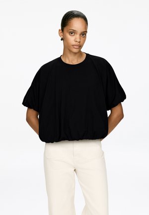 Schwarzes, kurzärmeliges Oberteil aus weichem Stoff mit gerafftem Saum. Die Silhouette ist locker geschnitten mit aufgeblähten Ärmeln. Kombiniert mit hellfarbigen Hosen.