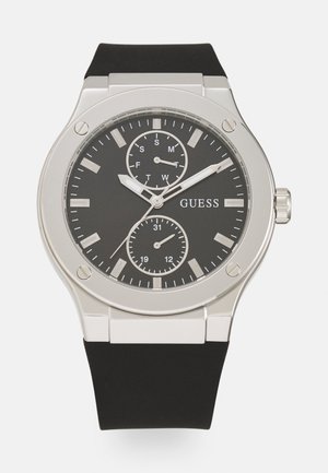 Silver case analog watch with black dial, silver hour markers, two subdials displaying day of week and 24-hour time, and black strap.