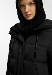 Vrouw die een zwarte gebreide balaclava met capuchon en een zwarte gewatteerde pufferjack met hoge kraag draagt, tegen een witte achtergrond.