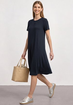 Femme marchant dans une robe noire au genou, baskets argentées, et portant un sac fourre-tout tissé beige avec une pochette en tissu à l'intérieur.